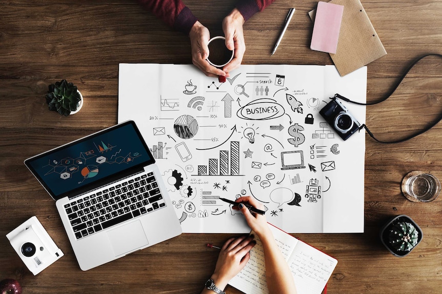 Overhead shot of a laptop, business plan, camera and hands around a coffee mug.