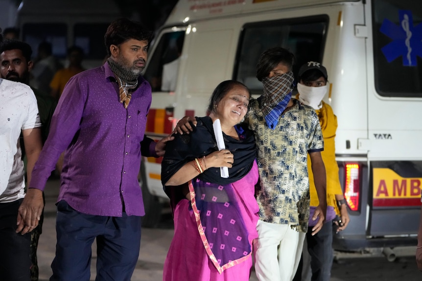 A woman cries out, while being held up by a man beside her, standing in front of an ambulance