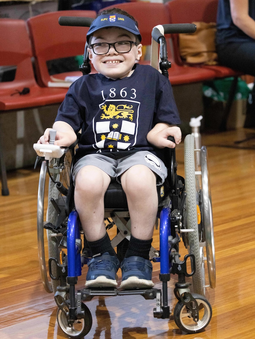 An 11-year-old boy in a wheelchair wearing a Sydney University t-shirt smiles.