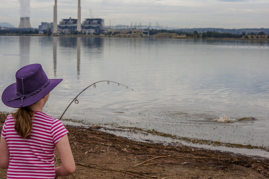A young girl reels in a fish at a lake near a coal-fired power station.