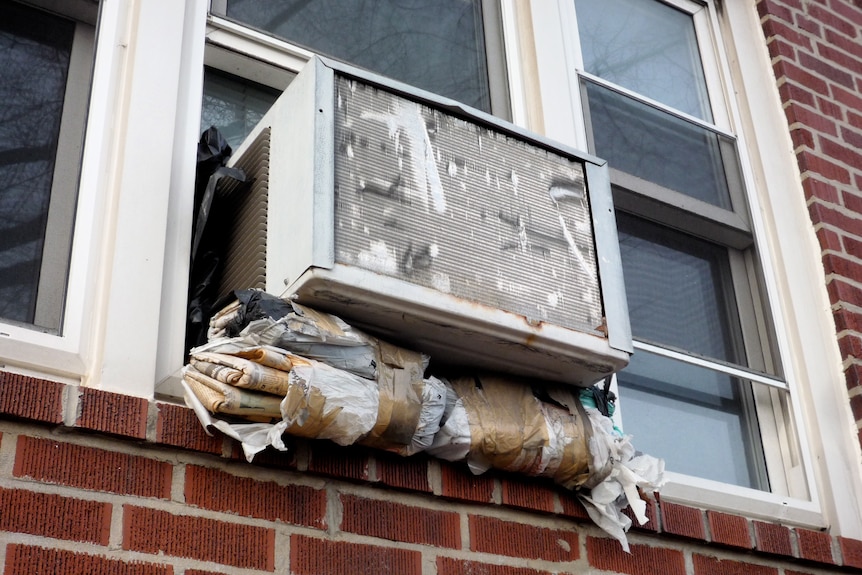 An aged box air con hanging out a window, propped up with newspapers.