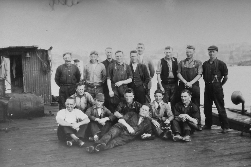 A black and white image of young men - nine stand in a row with six sitting in front of them with water behind them. 