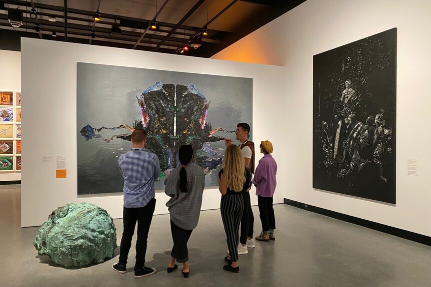 Five people stand with their backs to the camera looking at a large painting which is styled like psychologist's Rorschach test