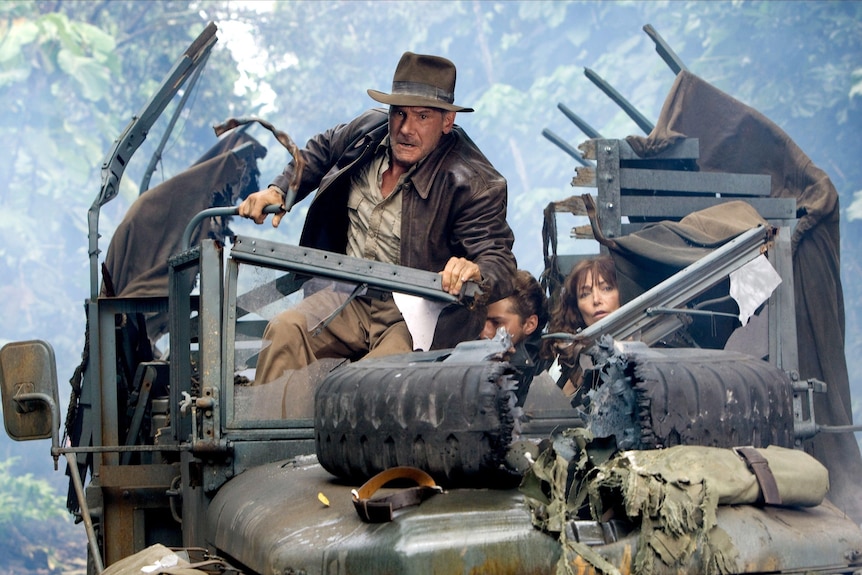 Indiana Jones stands atop a damaged jeep in a jungle.
