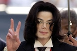 Michael Jackson makes a 'peace' gesture with one hand. Someone is holding an umbrella above his head
