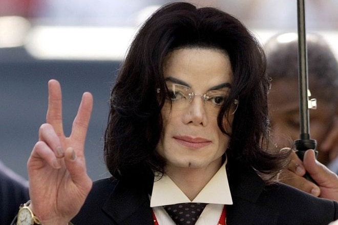 Michael Jackson makes a 'peace' gesture with one hand. Someone is holding an umbrella above his head