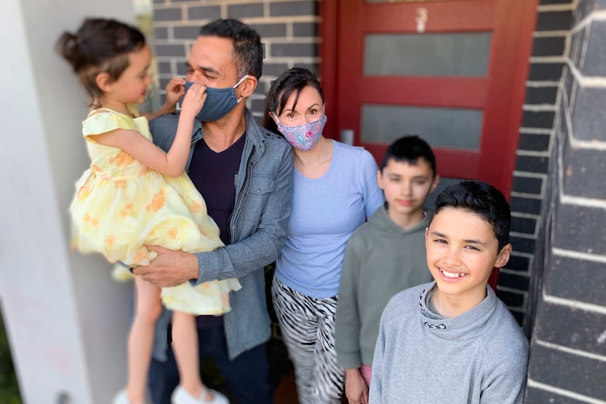 A girl adjusts the face mask of her father as her mother and two brothers pose outside the front door of their home.