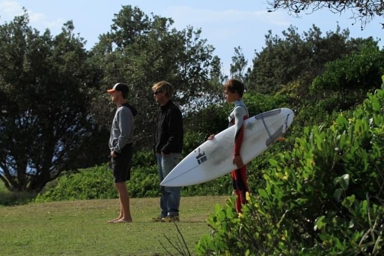 three men standing on grass looking out, one holding a surfboard