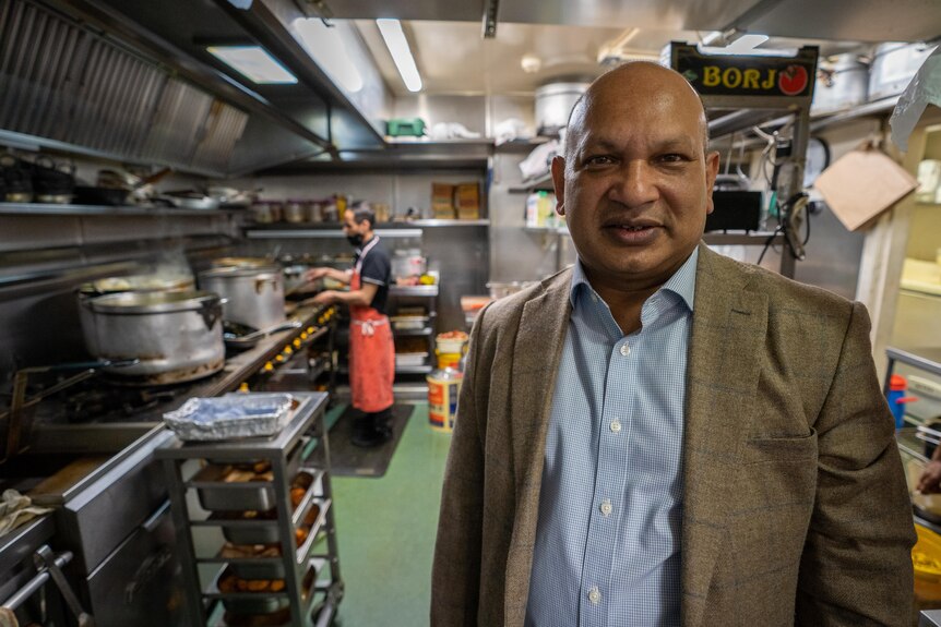 A bald man in a suit stands in a restaurant kitchen.