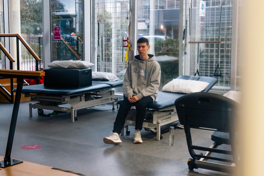 A teenage boy sits on the end of a bed in a hospital setting.