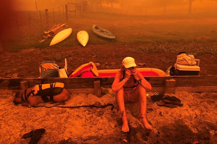 A woman sits with her head in her hands alongside a resting dog during bushfires at Fisherman's Paradise.
