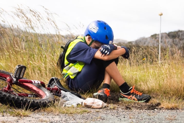 A boy sits next to a unicycle, by the side of a road, with his head bent down on his knees.