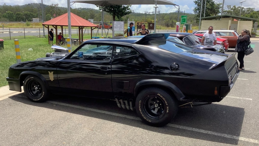 A black car with a spoiler on the back and parts of the engine protruding from its bonnet in a car park