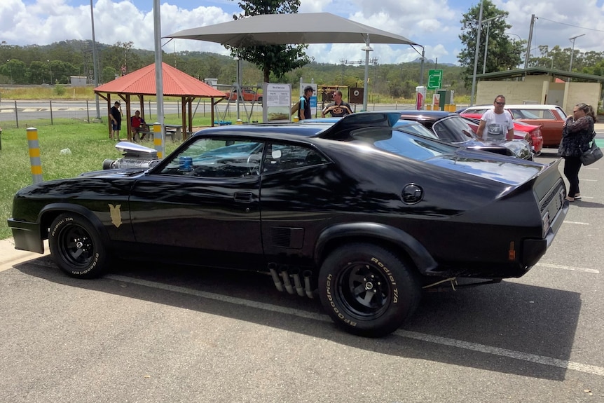 A black car with a spoiler on the back and parts of the engine protruding from its bonnet in a car park