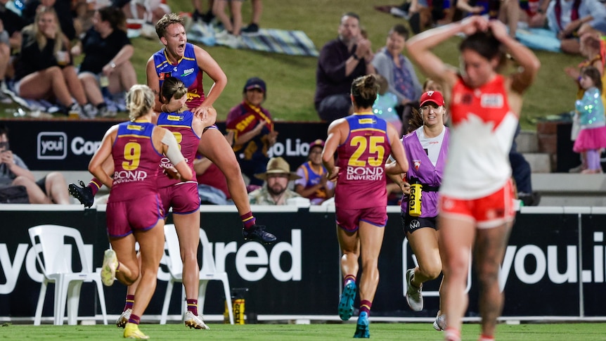 A Brisbane Lions AFLW player roars in celebration as a teammate lifts her in the air after a goal.