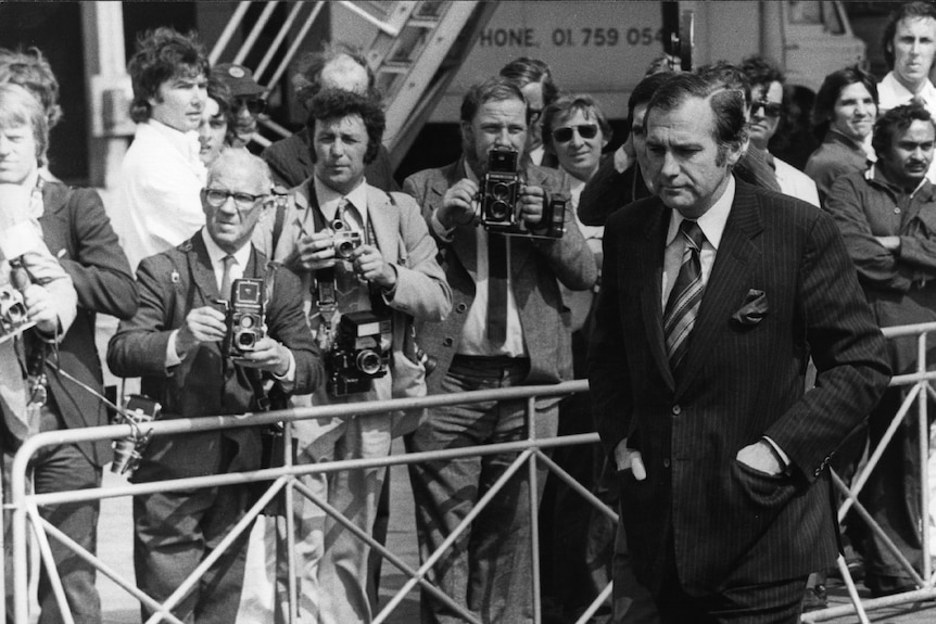 A black and white photo of a man, his hands in the pockets of a dark suit, walking in front of a pack of photographers