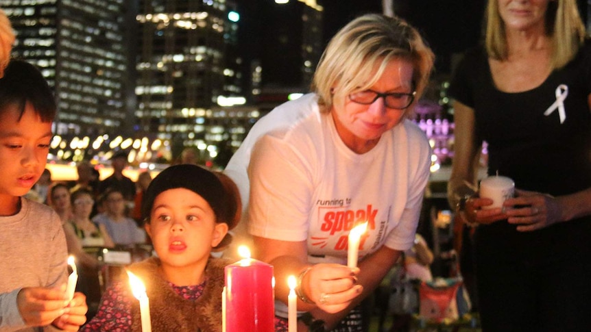 Rosie Batty holds a candle at a table near children who are also lighting candles, the lights of Brisbane CBD in the background
