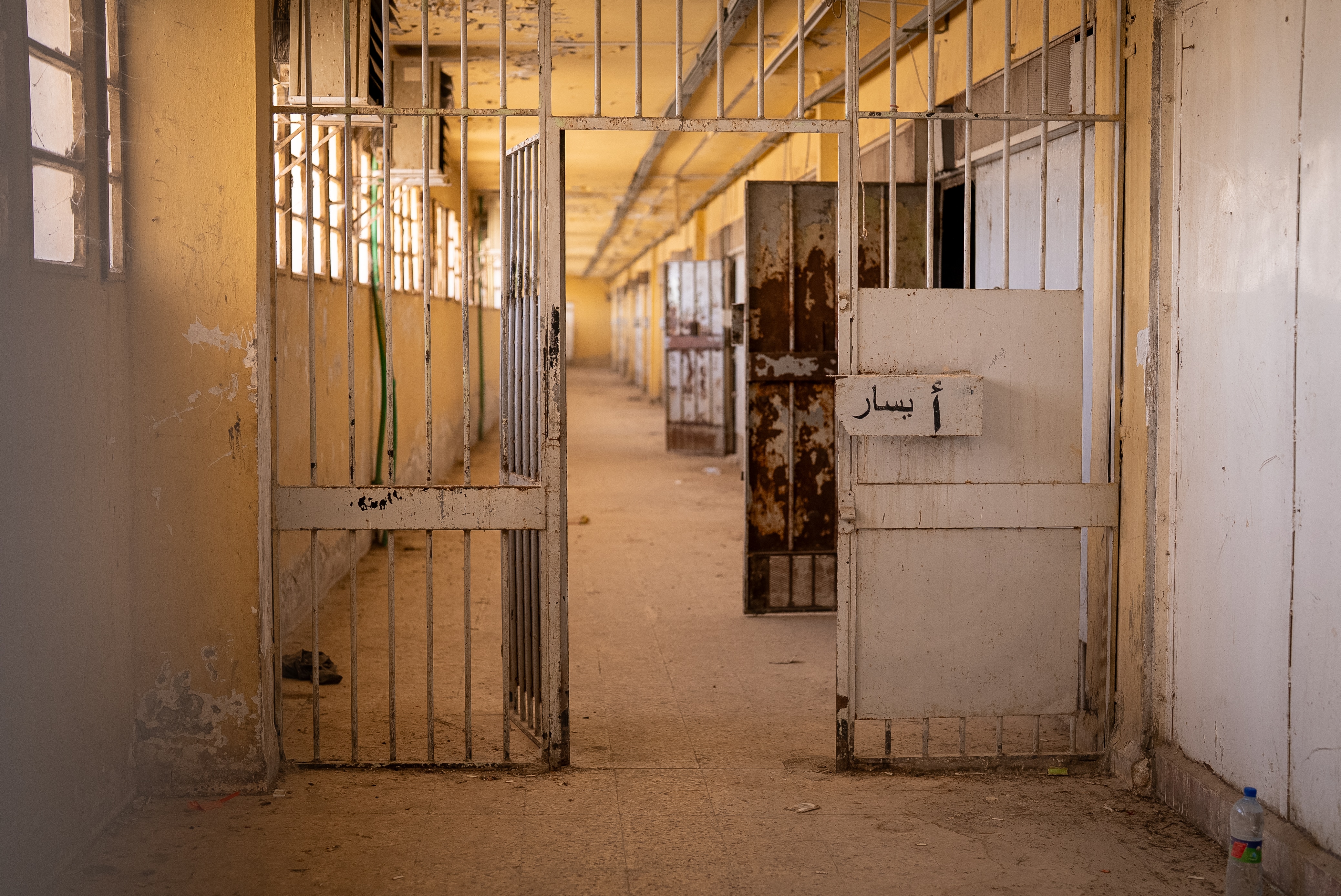 A prison hallway with unlocked gates and doors. The walls are yellow and We find windows to the left.