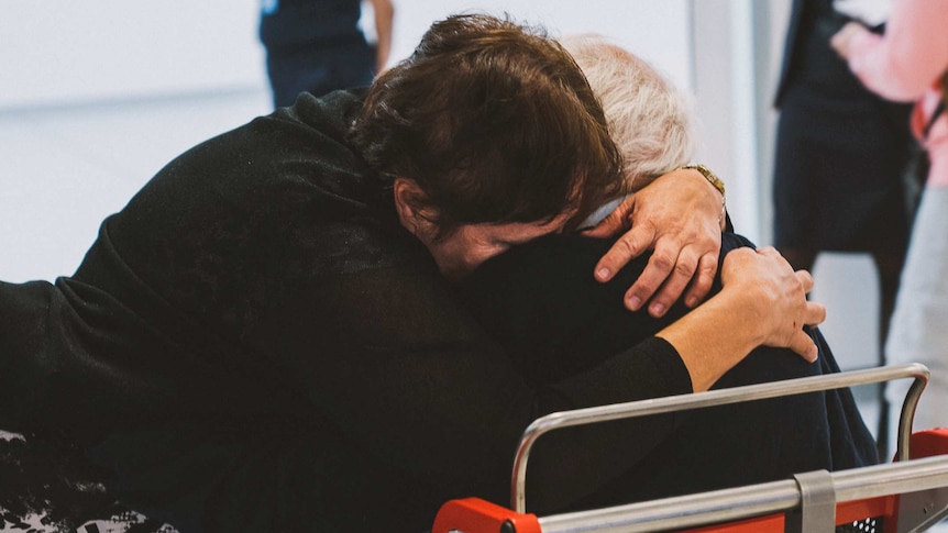 A woman in a black top and black and white dress hugs an elderly man in a wheelchair.