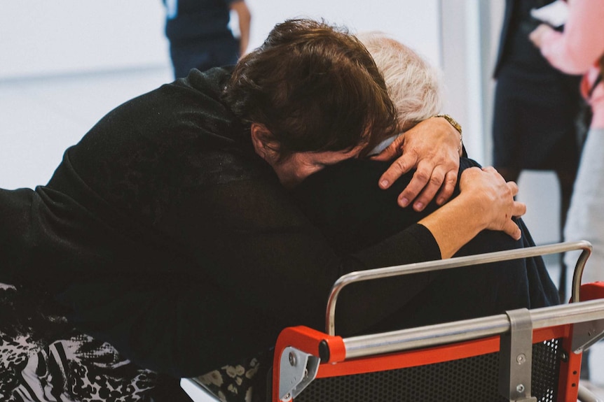 A woman in a black top and black and white dress hugs an elderly man in a wheelchair.