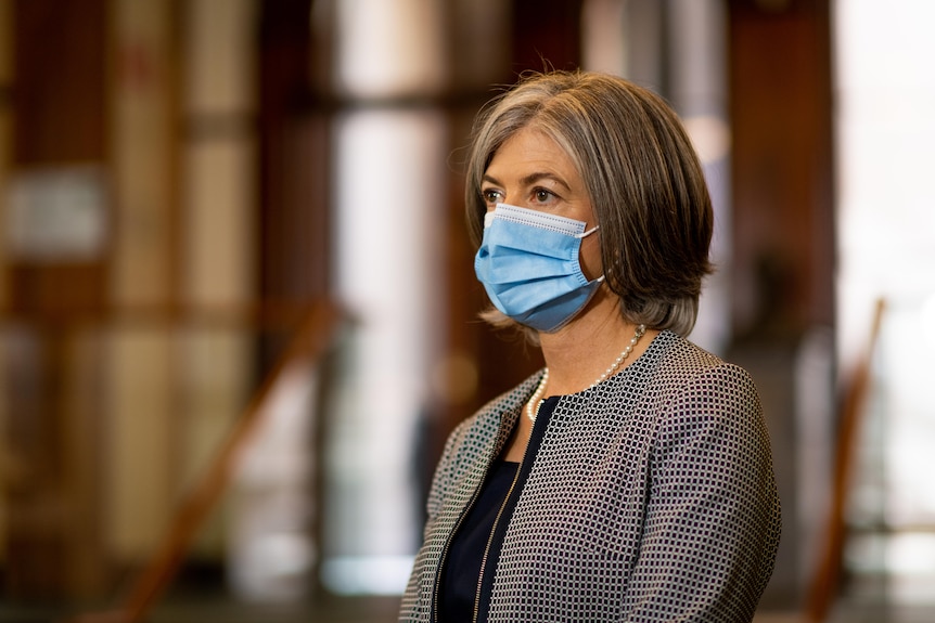 A woman stands side-on looking straight ahead, wearing a face mask