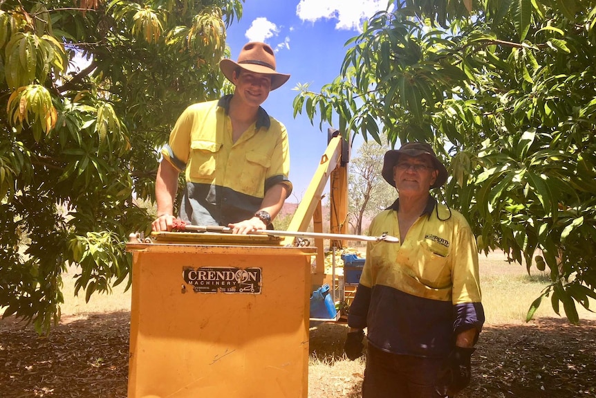 Two volunteers stand by a cherry picker in a mango orchard.