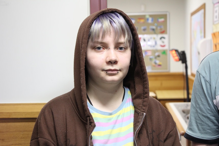 A student wearing a brown hoodie and a rainbow-pattern shirt stands inside, looking at the camera.