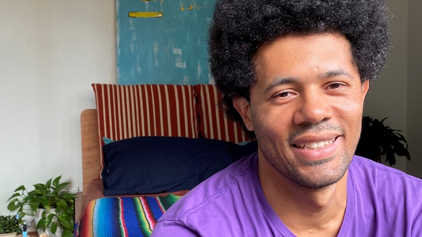 A smiling man sitting at the end of a bed wearing a purple t-shirt