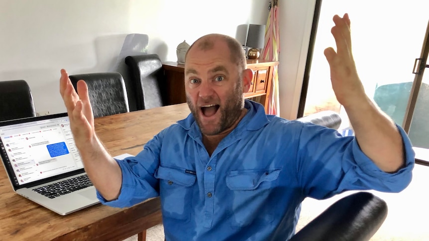 A man sits at a timber desk and laptop, with his hands in the air and a look of great surprise on his face. 