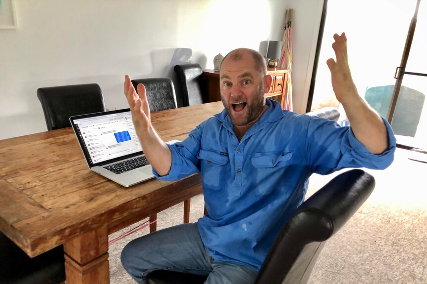 A man sits at a timber desk and laptop, with his hands in the air and a look of great surprise on his face. 