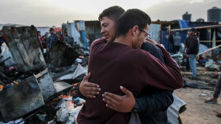 Two men hug each other crying in front of the burnt out wreck of a tent camp