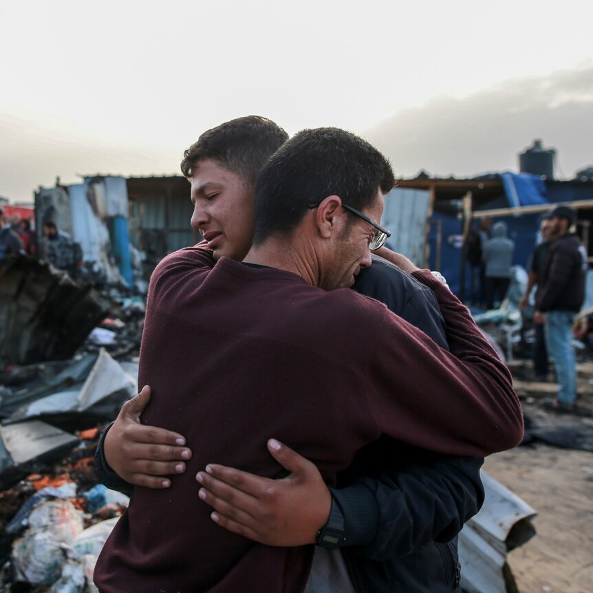 Two men hug each other crying in front of the burnt out wreck of a tent camp