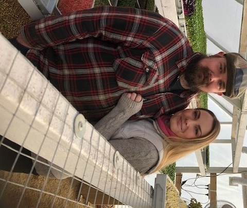 A young man and woman wearing warm clothes stand together in a greenhouse in front of trays of microgreens.