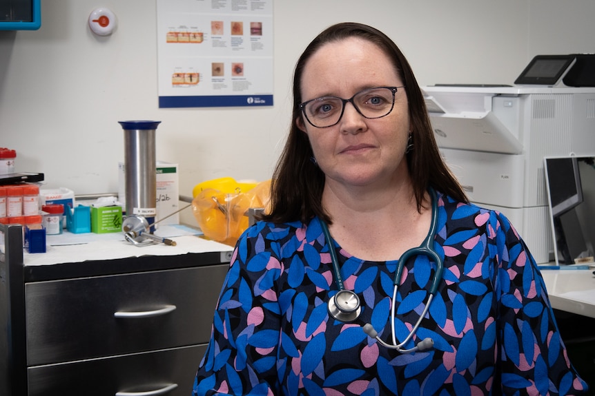 A woman in a blue shirt with a stethoscope around her neck sits in a doctors surgery. 