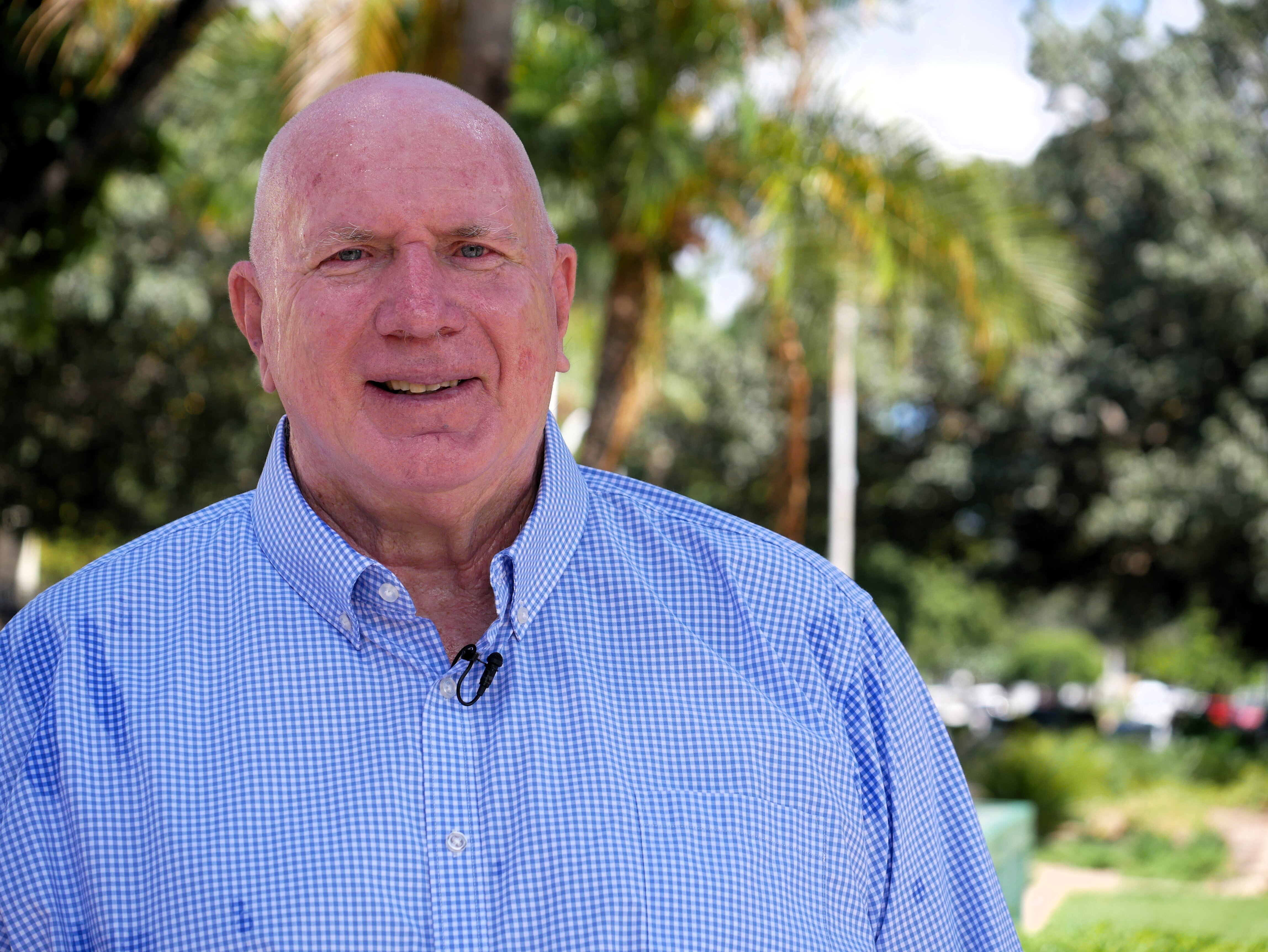 Um homem careca de meia-idade, com uma camisa xadrez azul, sorri para a câmera em um parque.