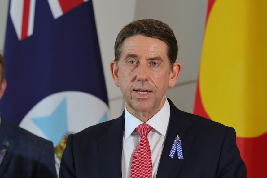 A dark-haired man in a dark suit stands in front of some flags and speaks to the media.