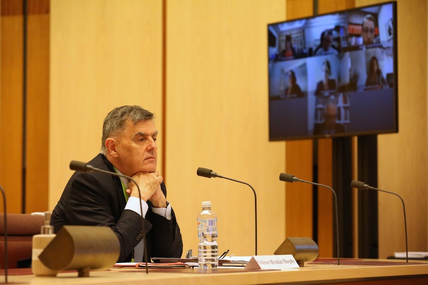 A man sitting at a desk in front microphones rests his chin on his hands with a tv screen and people on Skype in the background.