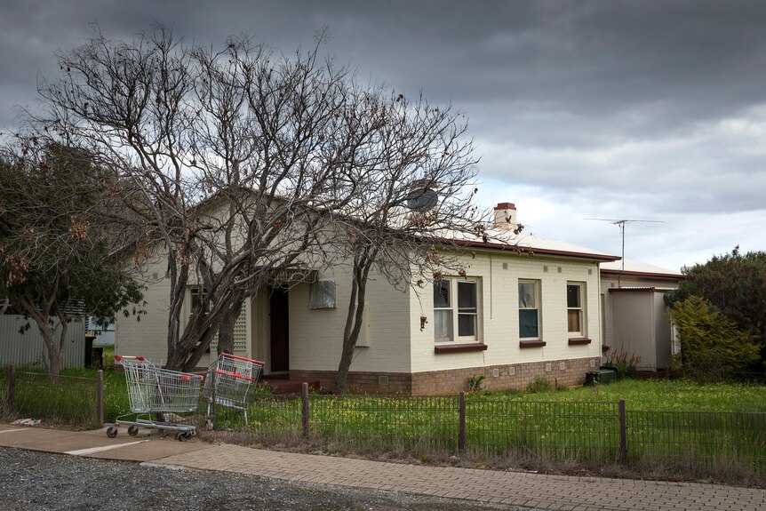 Colour photo of a cement home with grass front yard and wire fence, with two supermarket trolleys out the front of it.