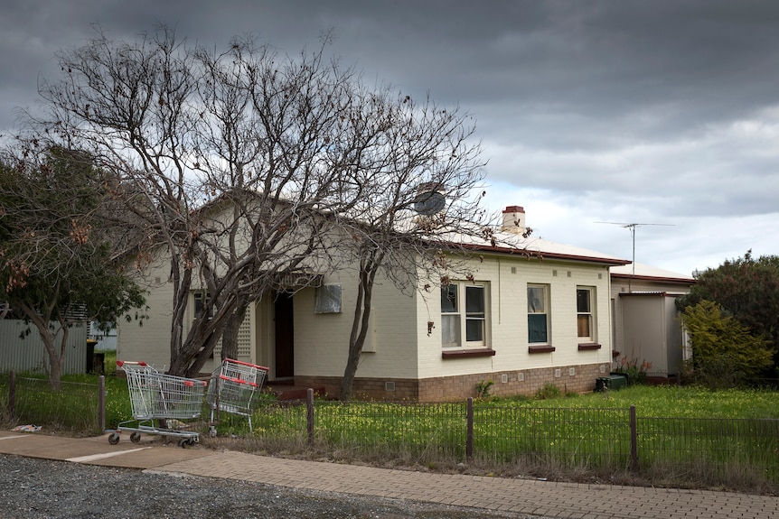 Colour photo of a cement home with grass front yard and wire fence, with two supermarket trolleys out the front of it.
