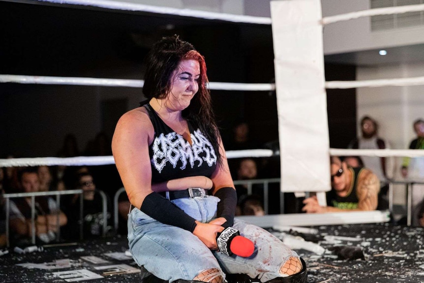 A sweating woman rests on the floor on her knees in a boxing ring, holding a microphone that leans on her knee and smiling.