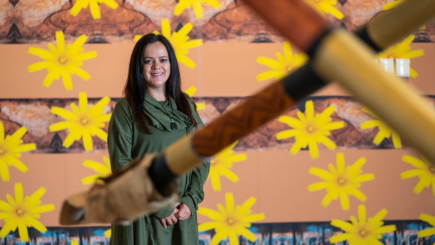 Wurundjeri woman with long dark hair wears olive green dress in front of mixed media artwork featuring bright yellow flowers.