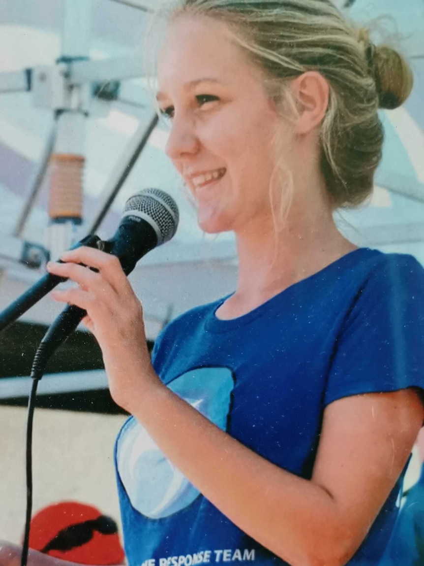 Old-look photo of smiling blonde teenager in blue shirt giving a speech, holding a microphone and paper notes in the other hand.