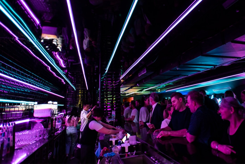 A crowded bar with neon lights and staff serving drinks to customers.