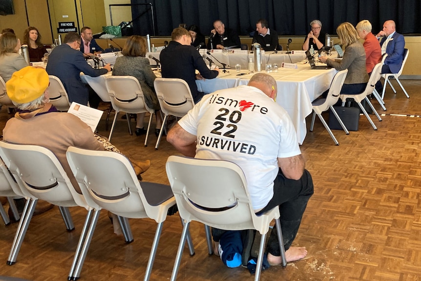 A man with an "I Survived Lismore 2022" shirt at a flood meeting inquiry.