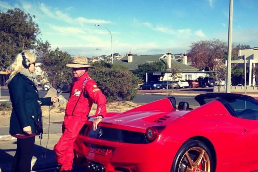A man in all red leans on his Ferrari and chats to a reporter. 