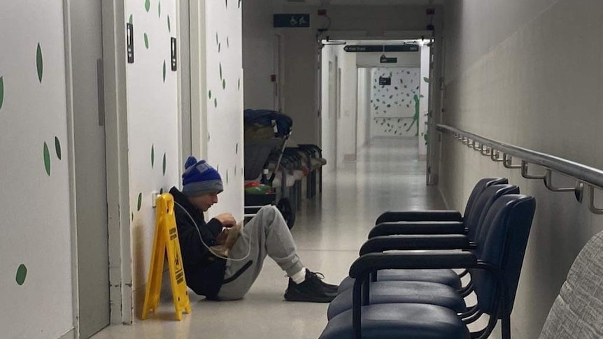 A teenager wearing a beanie, hoodie and track pants, sits against a wall in a hospital  while charging his mobile phone