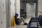 A teenager wearing a beanie, hoodie and track pants, sits against a wall in a hospital  while charging his mobile phone