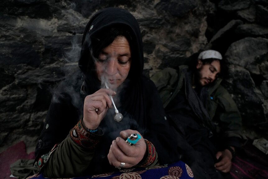 A middle-aged Afghan woman with a nose ring and rich-looking clothing smokes heroin from a small white glass pipe.