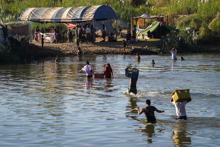People cross a river carrying large boxes over their heads and between two people. On the other bank sit makeshift tents