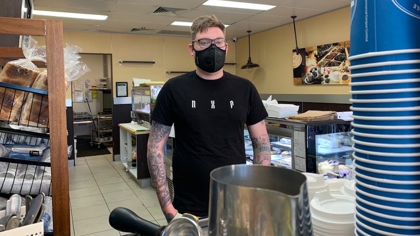 A man dark clothes, wearing a face mask, stands next to a coffee machine.